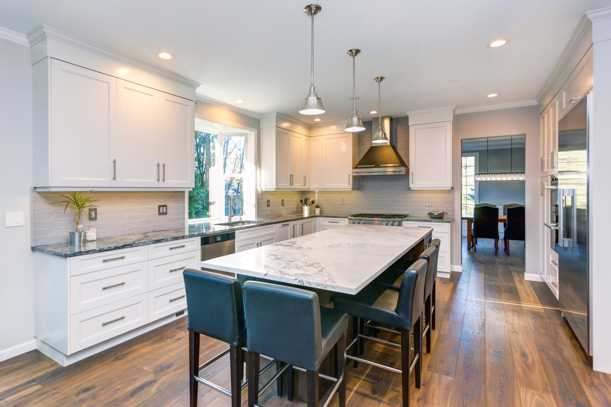 Luxury home interior boasts Beautiful black and white kitchen with custom white shaker cabinets, endless marble topped Kitchen Remodel Pricing Guide island with black leather stools over wide planked hardwood floor.